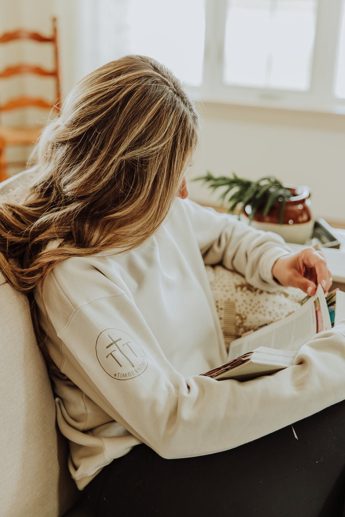 Person wearing a beige sweatshirt with a logo, sitting on a couch holding a book.